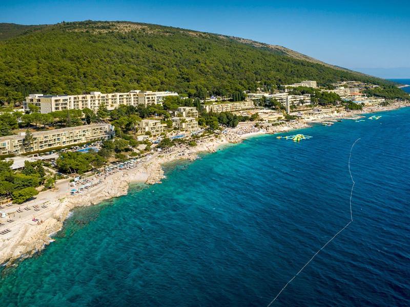 Línea costera con agua azul clara, playa y edificios frente a una colina boscosa al fondo.