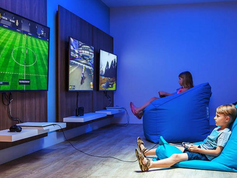 Dos personas juegan videojuegos en una sala moderna con pufs azules y pantallas grandes.