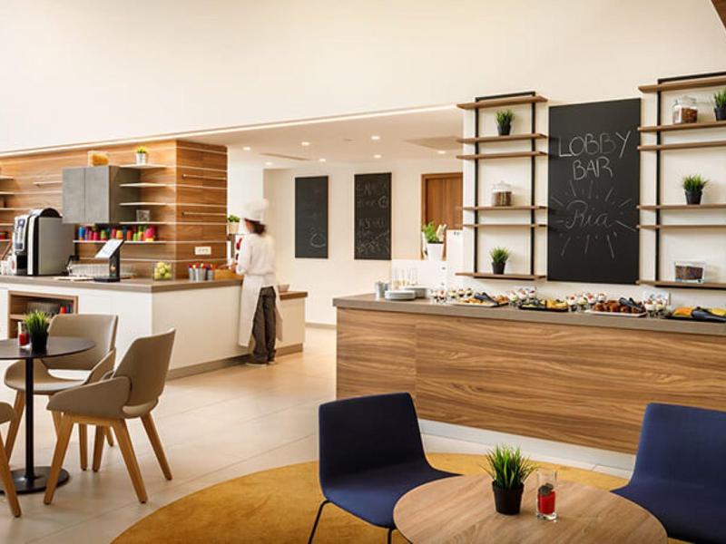 Lounge de hotel moderna con revestimiento de madera, mesas, sillas y barra de café al fondo.