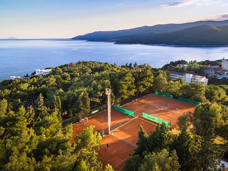 Paisaje con canchas de tenis, bosque y mar al fondo bajo cielo despejado.