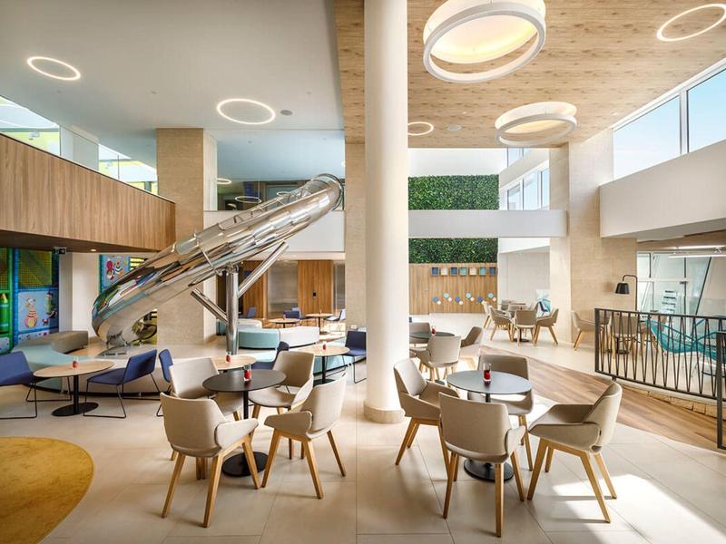 Área de salón moderna en hotel con sillas, mesas y un gran tobogán en espacio abierto.
