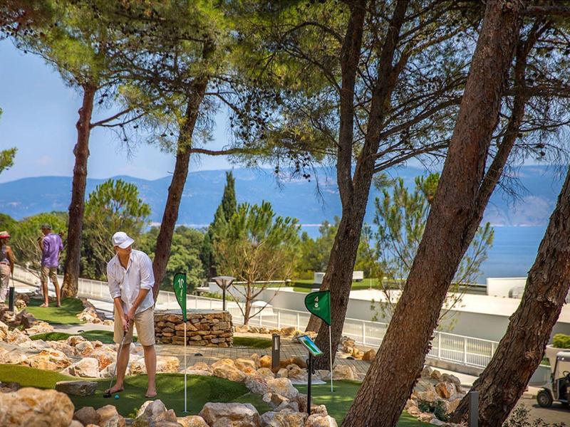 Hombre juega minigolf en un parque soleado y verde con vista al mar y árboles.