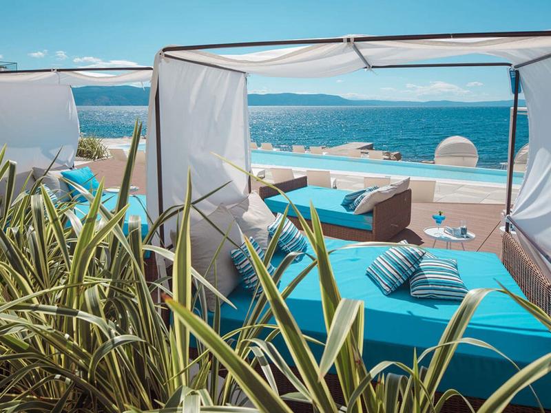 Vista de una tumbona acolchada azul bajo una cubierta blanca junto a una piscina con vista al mar.