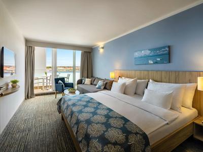 Modern hotelkamer met groot bed, bank, balkon en zeezicht bij daglicht.