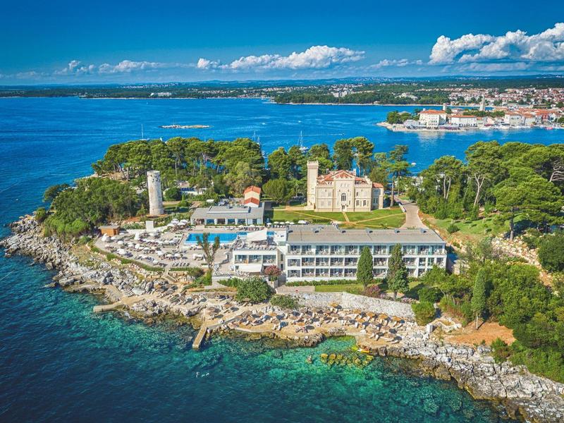 Hotelcomplex op een groen schiereiland met uitzicht op de blauwe zee en bewolkte lucht.