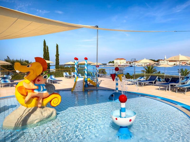 Kinderen spetteren in het ondiepe zwembad met kleurrijk speeltoestel en parasols op de achtergrond.