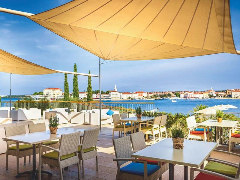 Terras met tafels, stoelen en zonneschermen met uitzicht op water en stad.