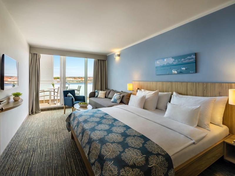 Modern hotelkamer met groot bed, bank, balkon en zeezicht bij daglicht.