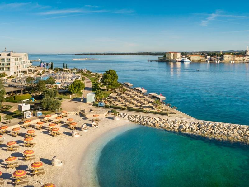 Strand met parasols en ligstoelen naast kalm blauw water en modern hotelcomplex.