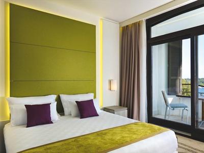 Modern hotel room with large bed, green wall panel, and balcony overlooking the sea.