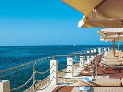 Promenadendeck am Meer mit weißen Pfeilern, Ketten und Sonnenschirmen bei klarem Himmel.