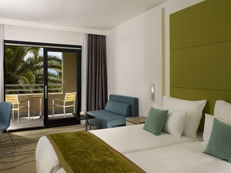 Modern hotel room with double bed, green headboard, and balcony overlooking palm trees.