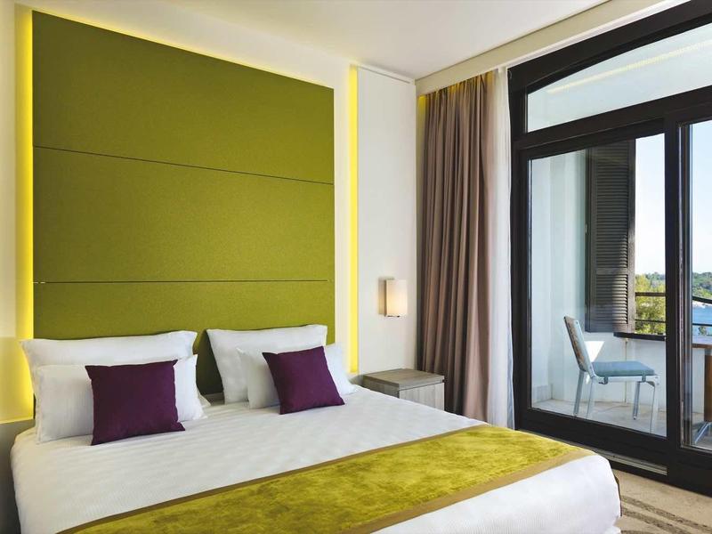 Modern hotel room with large bed, green wall panel, and balcony overlooking the sea.