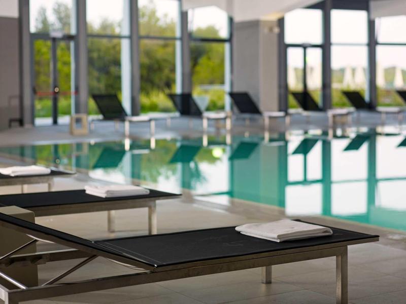 Modern loungers with white towels by a calm indoor pool with large windows.