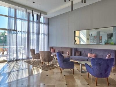 Espace lounge d'hôtel lumineux avec grandes fenêtres, fauteuils violets et miroir mural.