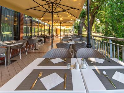 Terrasse avec tables dressées et parasols au bord d'une rivière, entourée d'arbres.