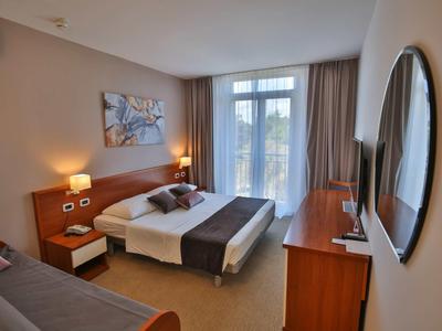 Světý hotelový pokoj s manželskou postelí, nočními stolky, pohovkou, velkým zrcadlem a výhledem na balkon.