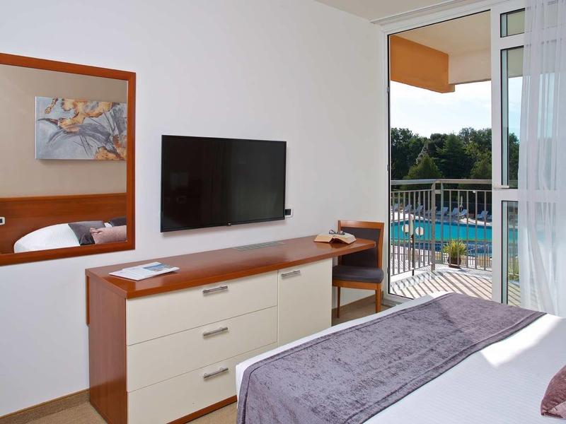 Moderní hotelový pokoj s TV, komodou a výhledem na balkon s bazénem.