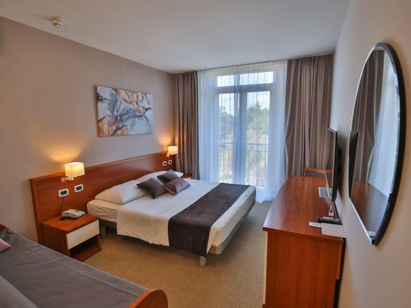 Světý hotelový pokoj s manželskou postelí, nočními stolky, pohovkou, velkým zrcadlem a výhledem na balkon.