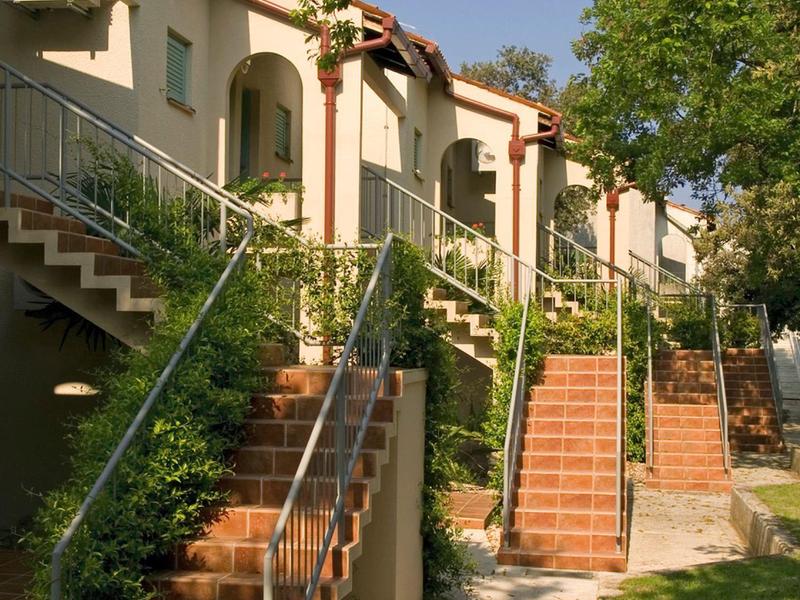 Řada hotelových pokojů s terasovými dveřmi, schody a zeleným okolím na slunci.