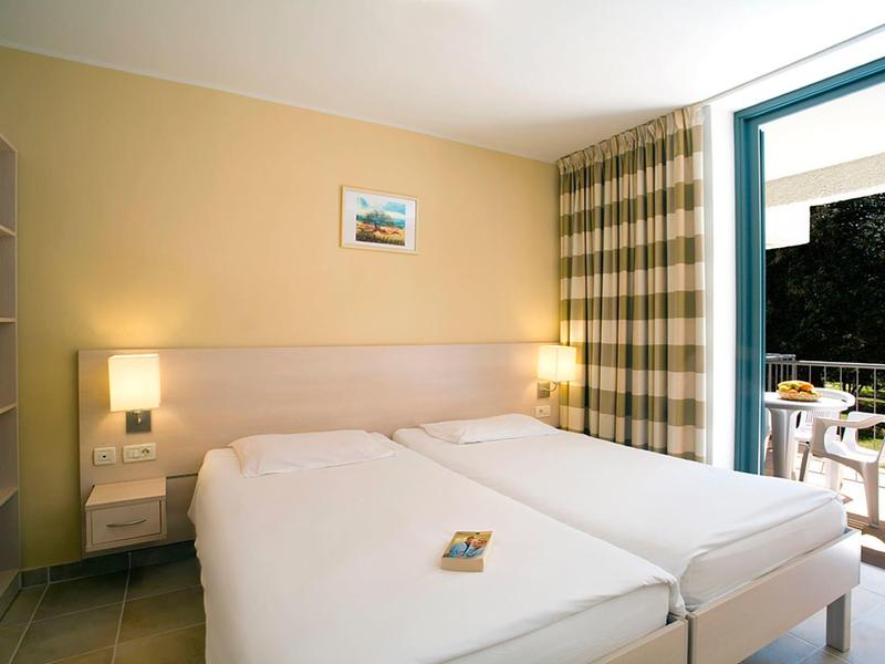 Prosto urządzony pokój hotelowy z dwoma pojedynczymi łóżkami i balkonem z widokiem na zieleń.