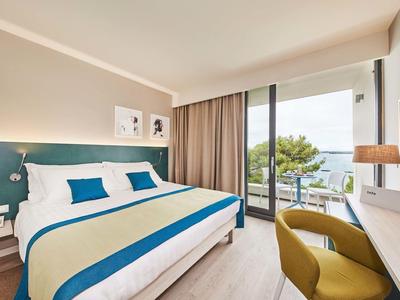 Habitación de hotel moderna con cama grande, escritorio y balcón con vistas al mar y árboles.