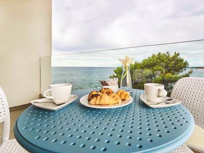 Mesa azul en balcón con café, cruasanes y vista al mar bajo cielo nublado.
