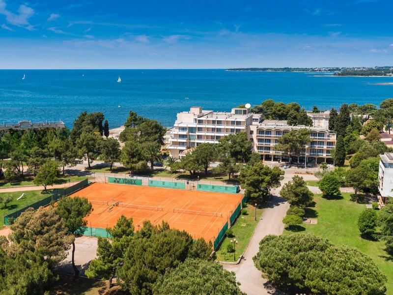 Blick auf ein Hotel mit Tennisplätzen, viel Grün und Meer im Hintergrund.