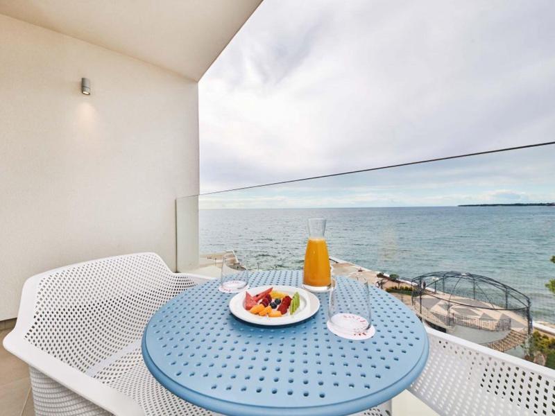 Balkon mit blauem Tisch, weißen Stühlen, Frühstück mit Saft, Obst und Marmelade, Meerblick, Glasgeländer.