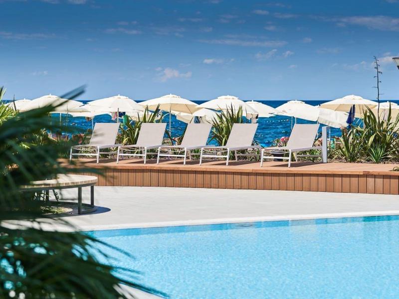 Blauer Pool mit Liegestühlen und Sonnenschirmen am Meer unter blauem Himmel.