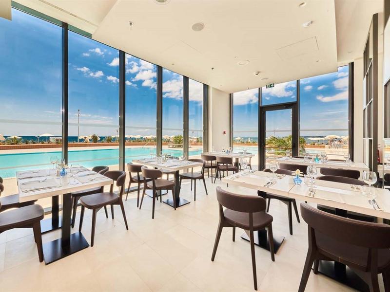 Modernes Restaurant mit großen Glasfenstern und Blick auf einen Außenpool und Himmel.