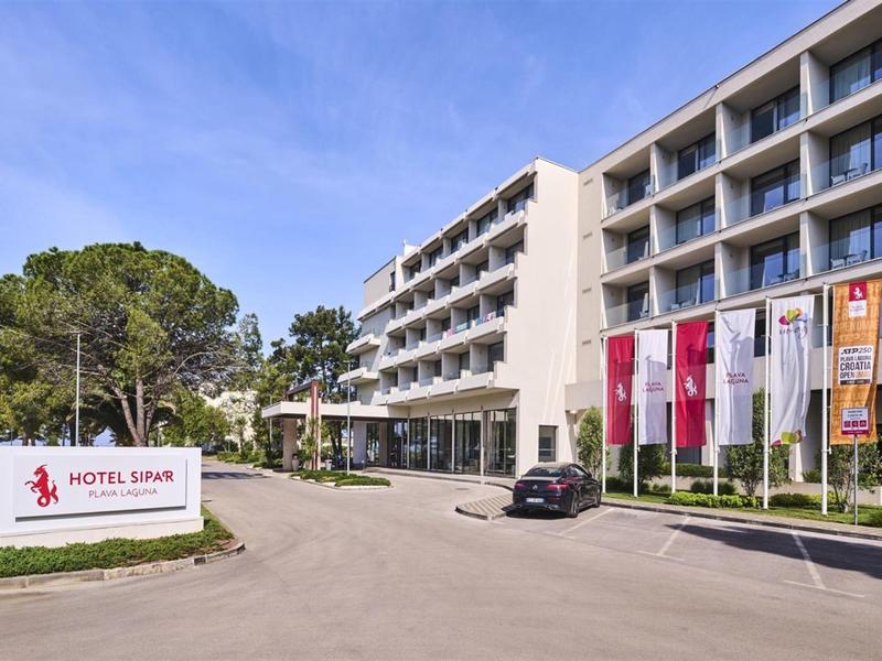 Modernes mehrstöckiges Hotelgebäude mit Fahnen, Parkplatz und grüner Umgebung unter blauem Himmel.
