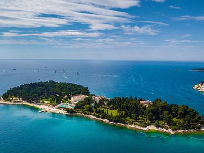 Grüne Insel mit Bäumen und Gebäuden, umgeben von blauem Meer unter bewölktem Himmel.