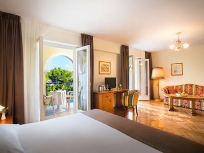Habitación de hotel luminosa con cama grande, balcón abierto, suelo de madera y zona de estar acogedora.