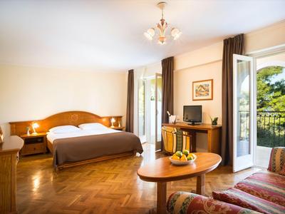 Habitación de hotel luminosa con suelo de madera, cama doble, zona de estar y balcón con vistas a árboles verdes.