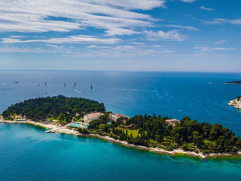 Green island with buildings surrounded by blue sea under cloudy sky.
