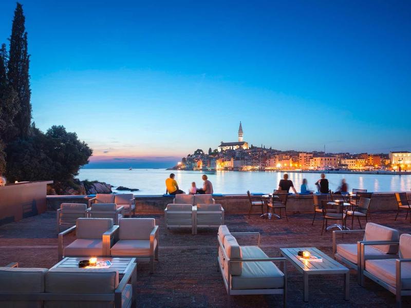 Evening terrace with seating areas overlooking the illuminated coastal town by the sea.