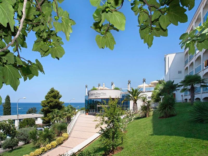 Vue d'un bâtiment hôtelier avec jardin et chemin menant à la mer sous un ciel clair.