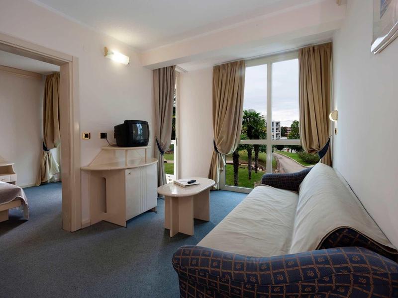 Chambre d'hôtel lumineuse avec canapé, télévision, minibar et vue sur un jardin verdoyant.