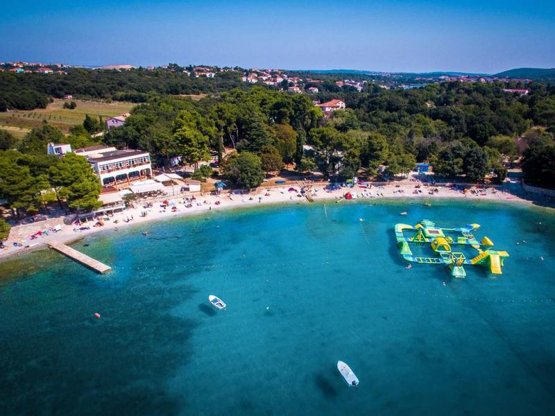 Klares blaues Meer mit Sandstrand, Wasserspielgerät, Booten und grünen Bäumen am Ufer.