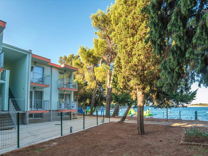 Mehrfamilienhaus mit Balkon, umgeben von Bäumen und Blick auf einen See bei klarem Himmel.