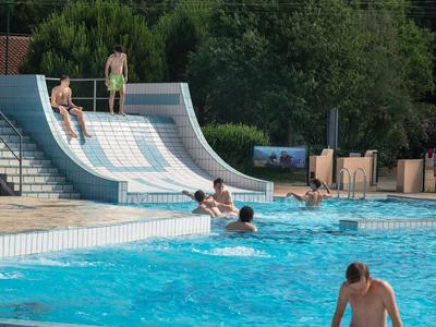 Basen z zjeżdżalnią wodną i ludźmi, otoczony drzewami w tle.