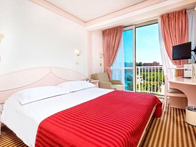 Helles Hotelzimmer mit Doppelbett, roter Bettdecke, Balkon und Meerblick.