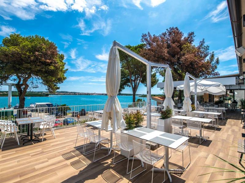 Taras nad morzem z białymi stołami, krzesłami i baldachimami pod błękitnym niebem.