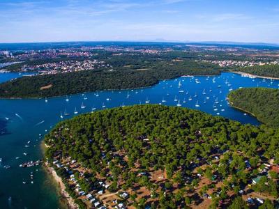 Luftaufnahme einer bewaldeten Halbinsel mit vielen Segelbooten im blauen Wasser und Häusern am Ufer.