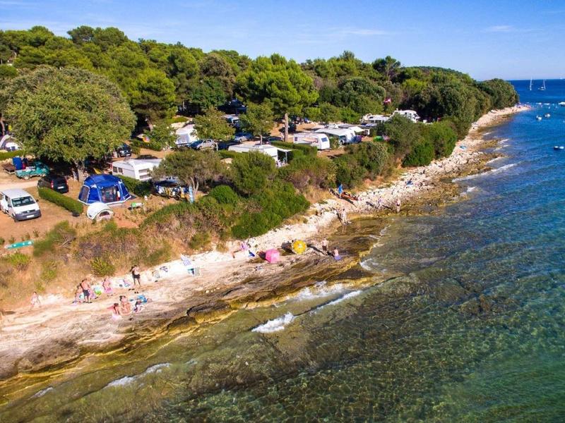 Küste mit klarem Wasser, Sandstrand, Bäumen und Campingfahrzeugen unter blauem Himmel.