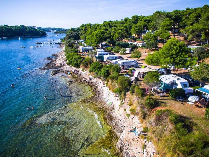 Küstenlinie mit klarem blauem Wasser, felsigem Ufer und mehreren Campingwagen zwischen Bäumen.