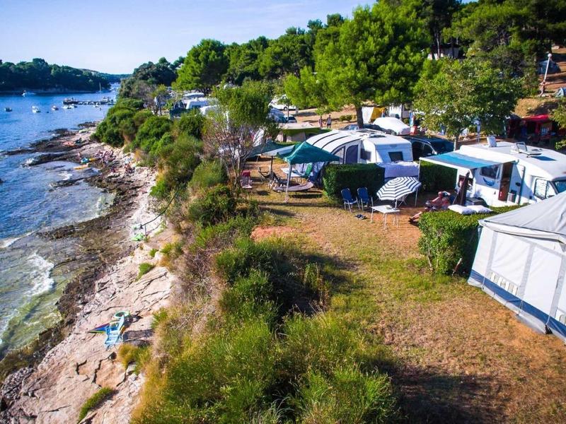 Campingplatz am Ufer mit Zelten, Wohnwagen, Kiesstrand und grünem Wald im Hintergrund.