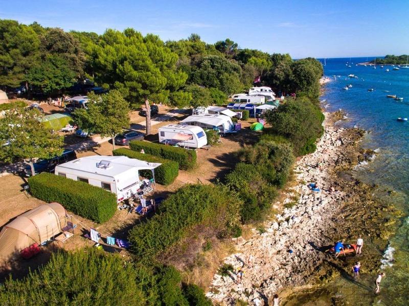 Campingplatz mit Zelten und Wohnwagen an bewaldeter Küste mit klarem blauem Wasser und Booten.