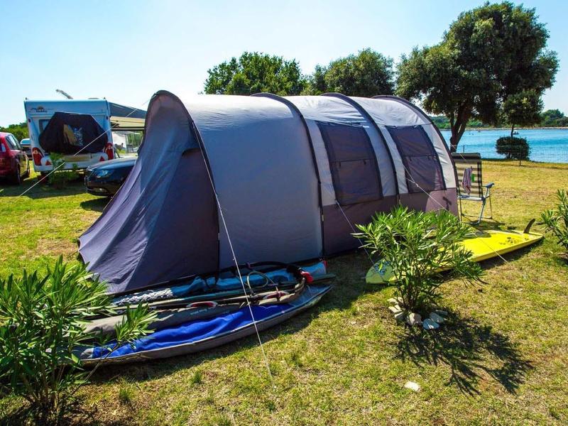 Großes graues Campingzelt auf grünem Gras mit zwei Surfboards und Bäumen im Hintergrund.